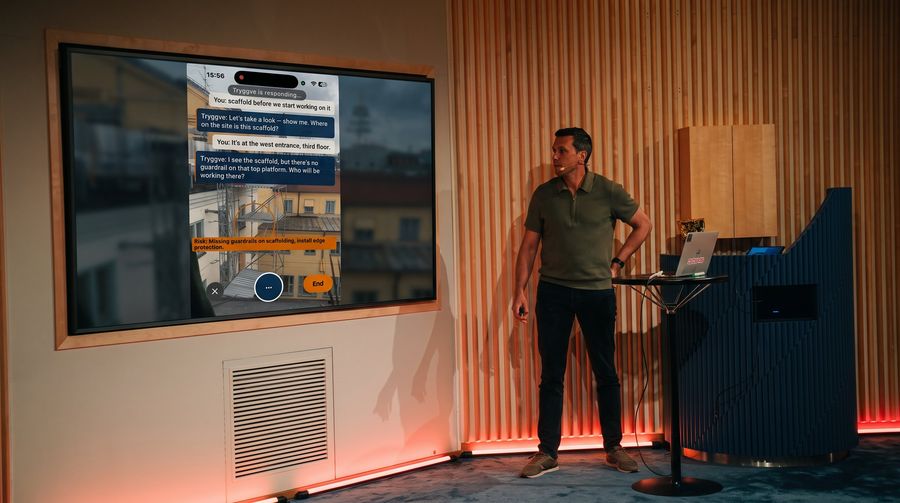 Robert Jansz pitchar Tryggve på scen i hackathonfinalen. Skärmen bakom honom visar ett livesamtal med Tryggve om en byggställning.